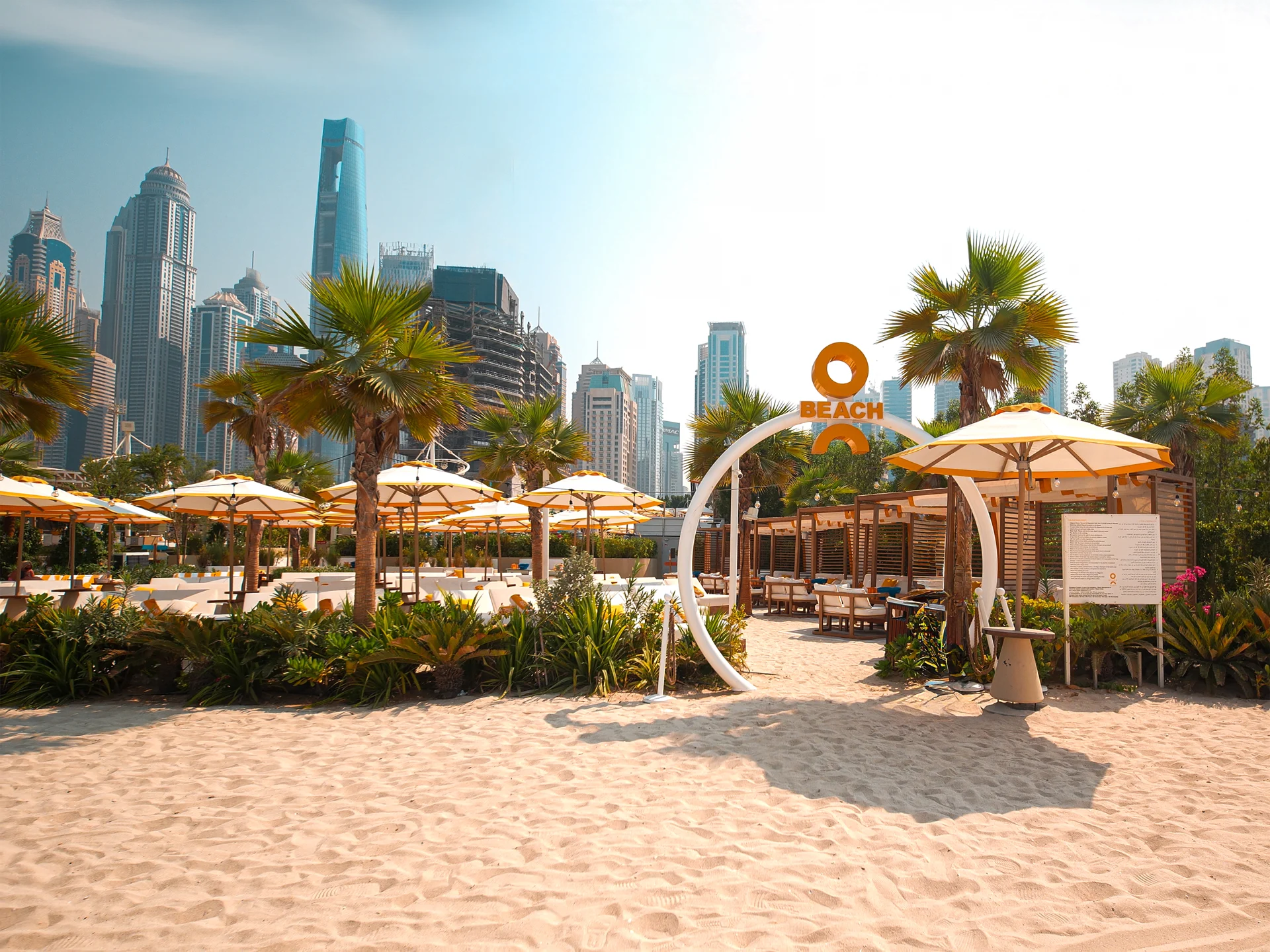 O Beach Dubai entry area with sand, palms, skyline, and brand sign.